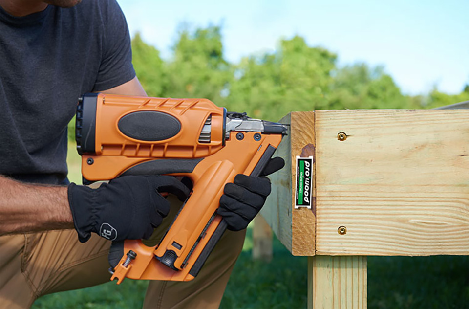 Gloved hands holding up a nail gun to lumber while building a deck